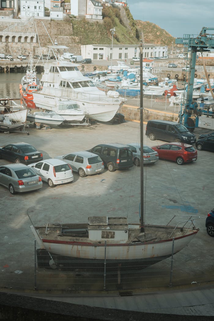 An old sailboat sits in a marina parking with yachts and cars on a sunny day.
